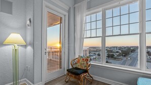 A room with a large window, a wicker chair, a floor lamp, and a view of a cityscape at sunset. - Beachfront Penthouse!  4Bd/3Bth (Virginia Beach)