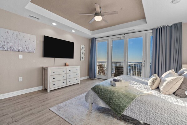 A bedroom with a large bed, a ceiling fan, a TV, a dresser, and a balcony with a view.