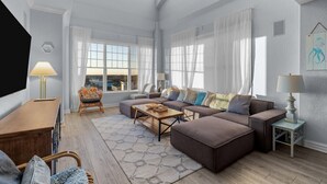 A spacious living room with a large sectional sofa, a wooden coffee table, and a patterned rug. There are two windows with sheer curtains, a wicker chair, and a small side table with a lamp. - Beachfront Penthouse!  4Bd/3Bth (Virginia Beach)