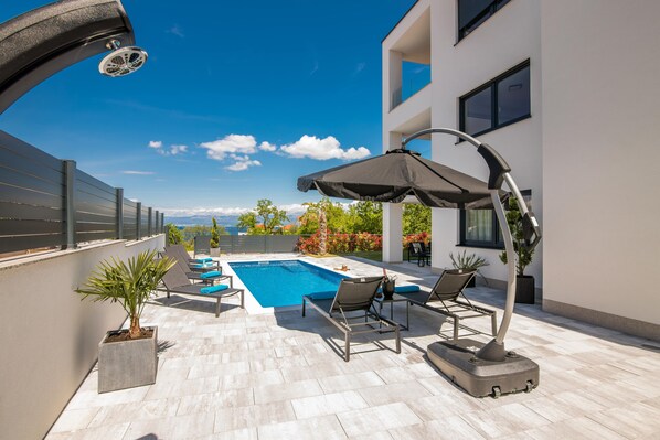 Relax by the pool surrounded by lounge chairs and a shaded area, with a view of the surrounding landscape from the adjacent building.