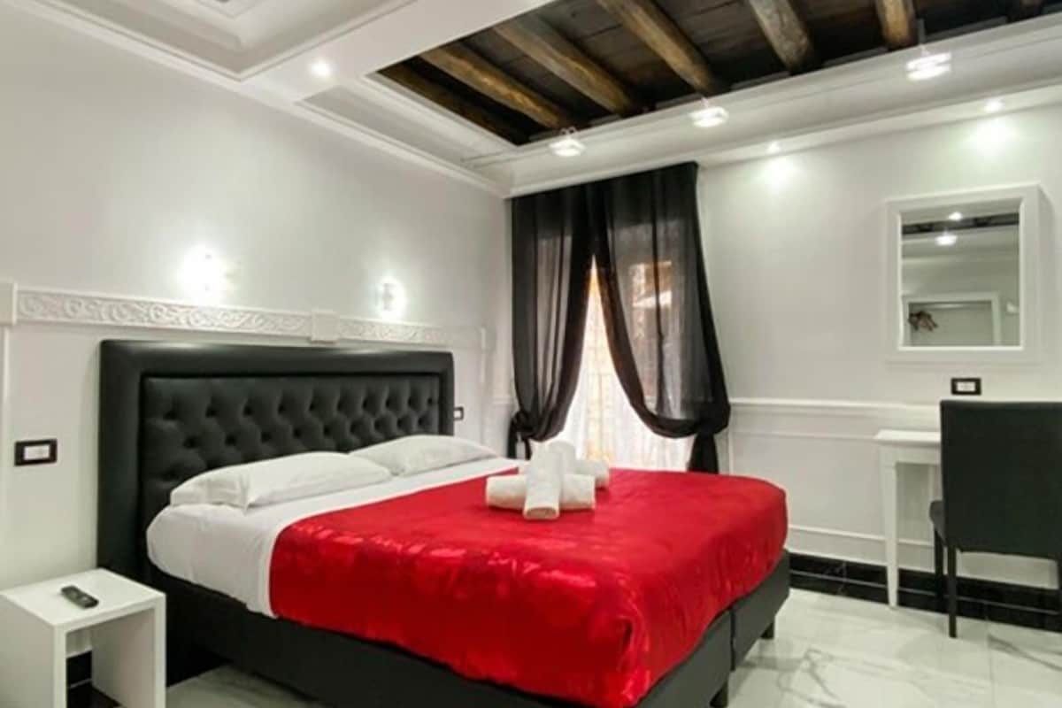 Een slaapkamer met een zwart bekleden hoofdeinde, een rode sprei, een witte nachtkastje en een klein bureau met stoel.