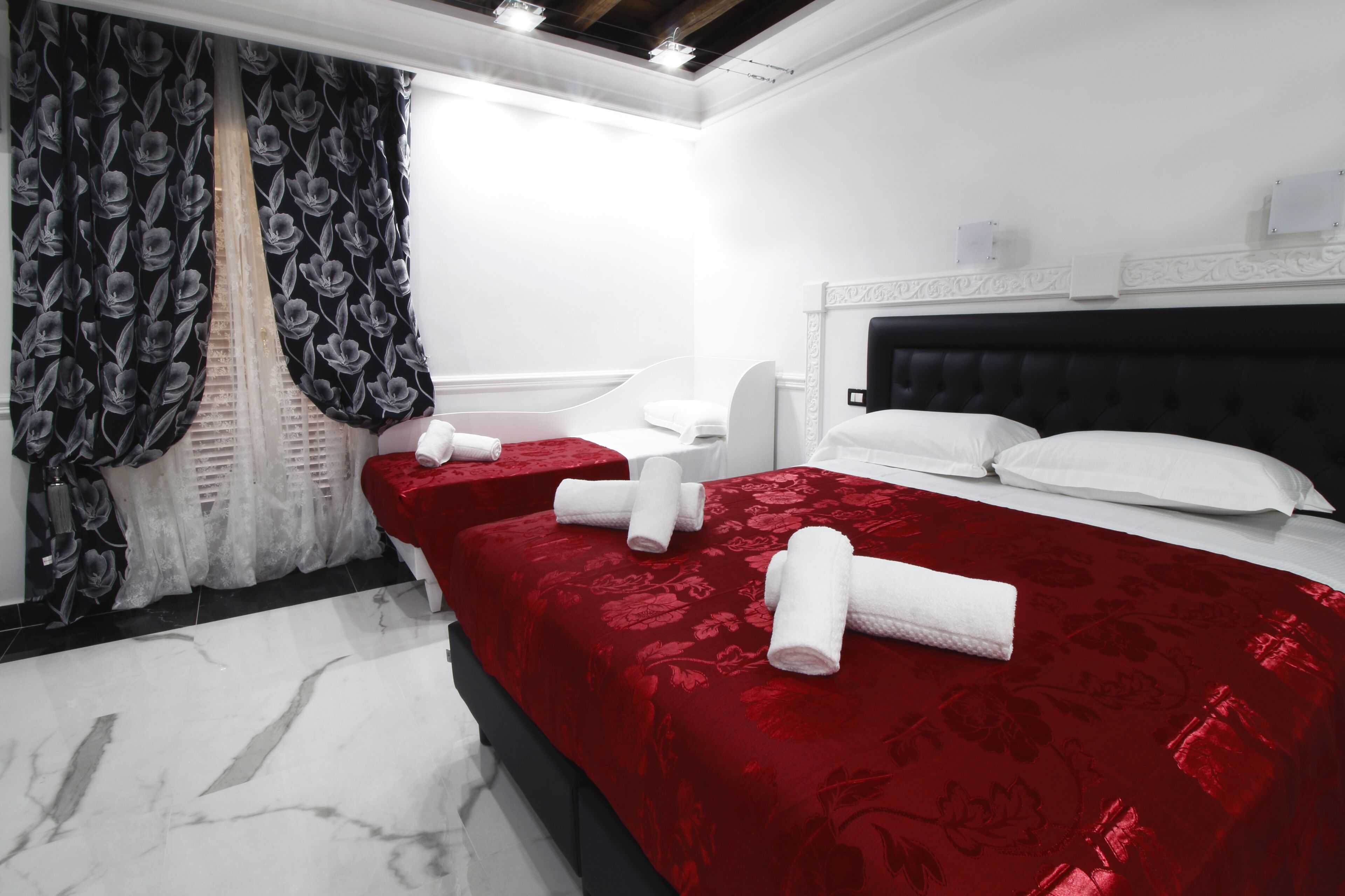 A hotel room with two beds, red bedding, white headboards, and a large window with black and white floral curtains.