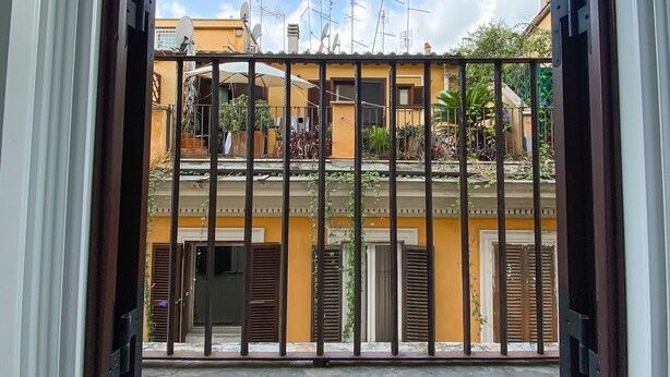Vista a través de una ventana con reja metálica, que muestra un edificio con balcones, antenas parabólicas y cielo despejado.