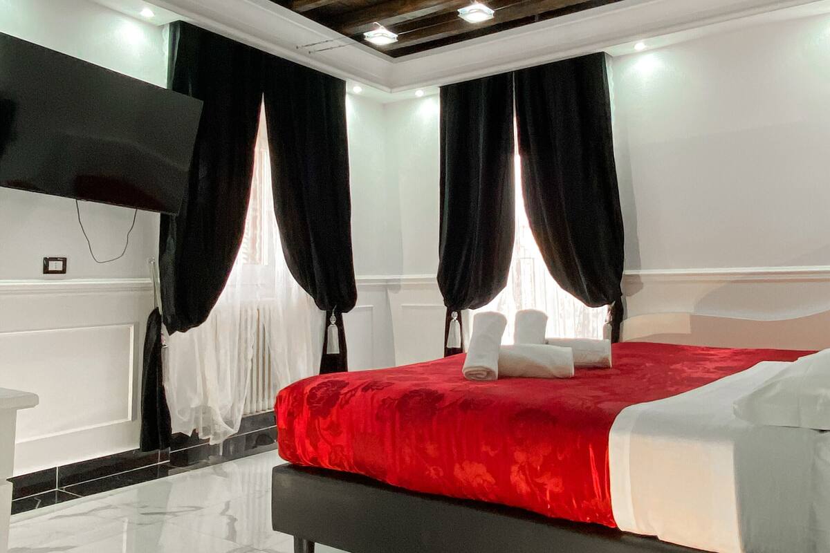 A modern hotel room with a large bed, dark wooden ceiling beams, white walls, and a marble floor.
