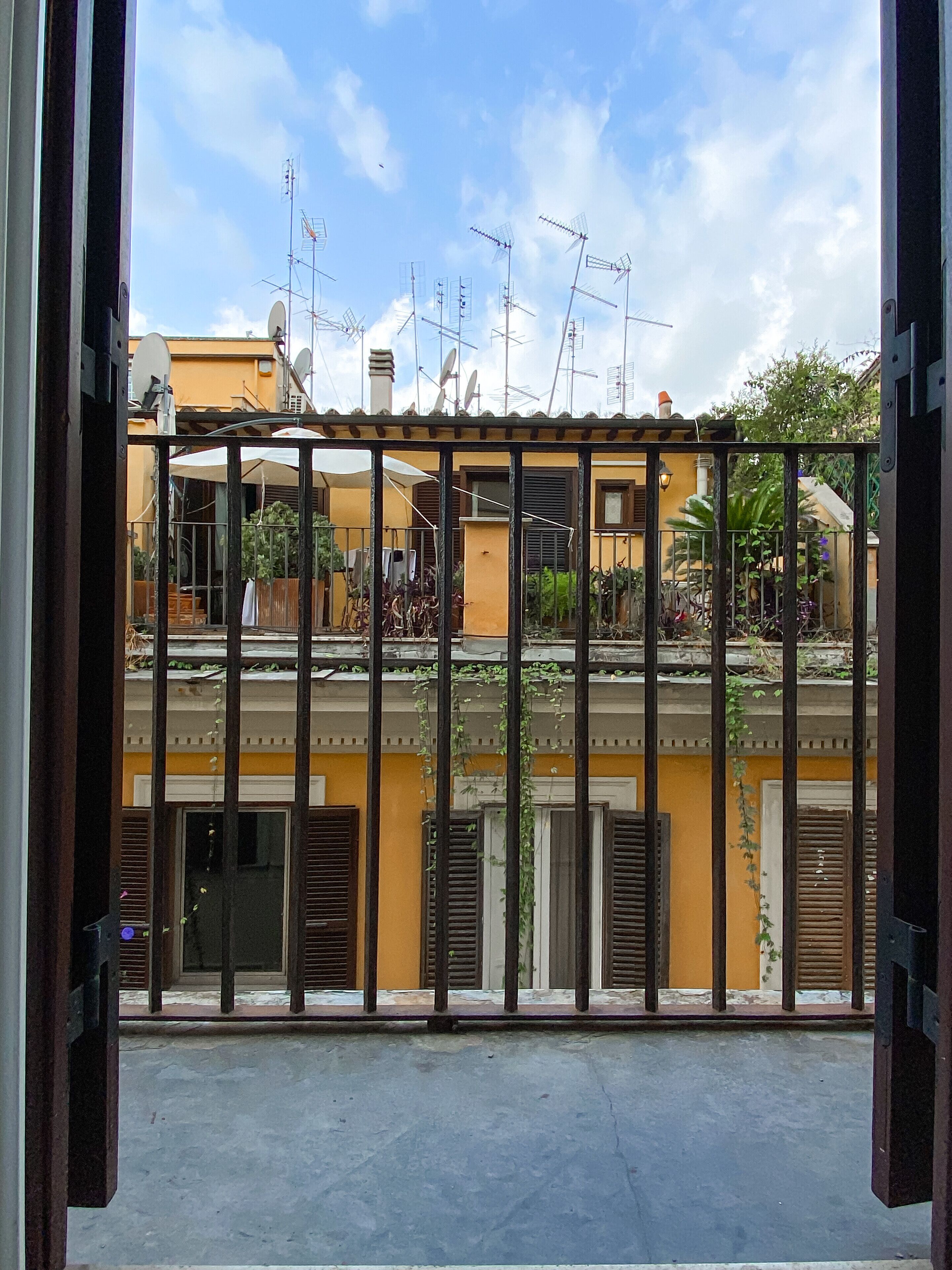A view through a metal gate of a building with multiple antennas, a yellow facade, and a balcony with plants.