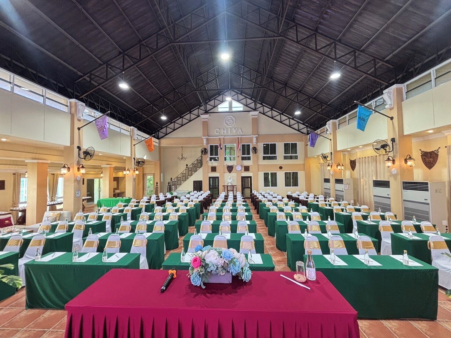 A large hall with rows of tables set for an event, a stage at the front, and a staircase leading to an upper level.