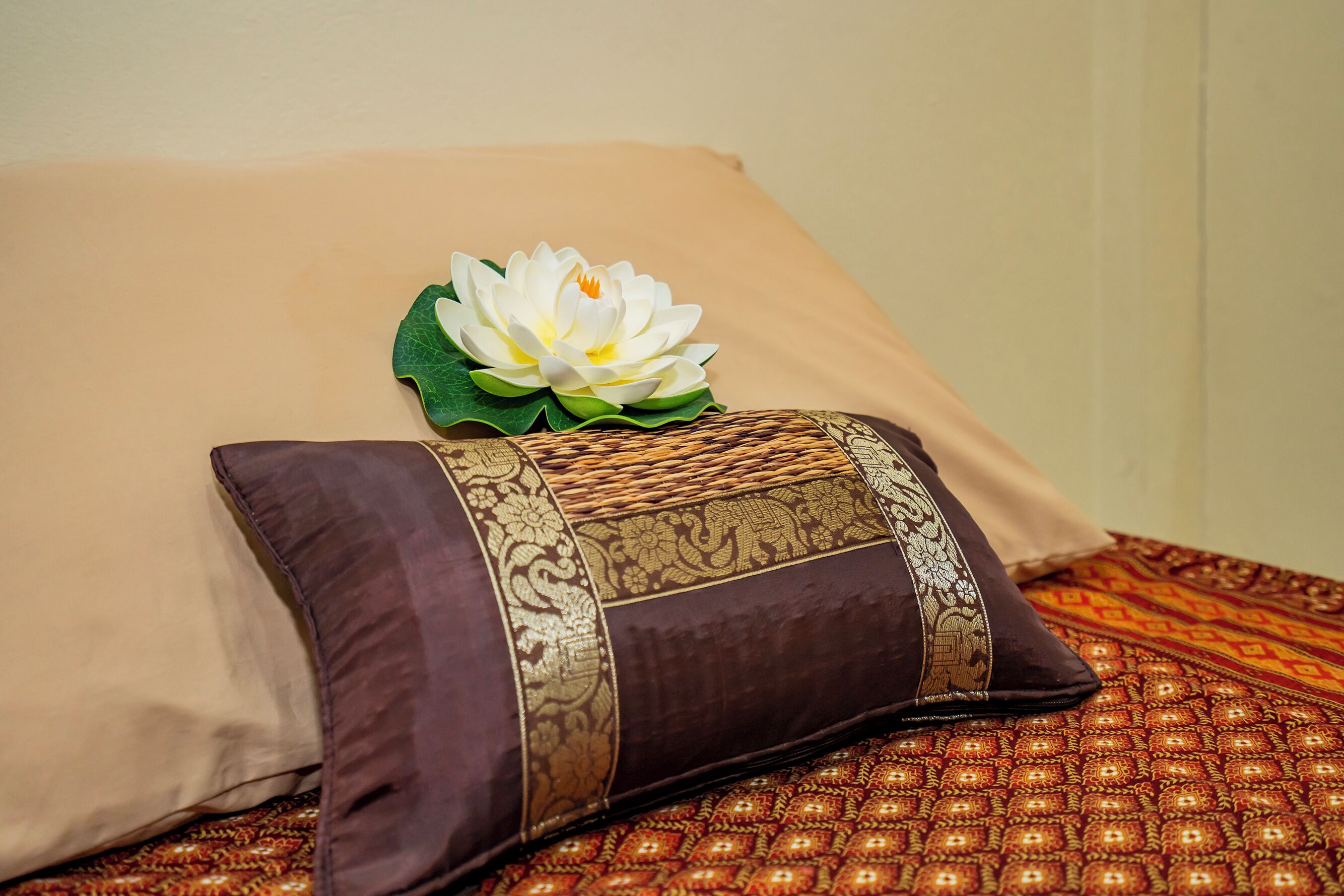 A bed with a beige pillow, a decorative pillow with a patterned design, and a white lotus flower.