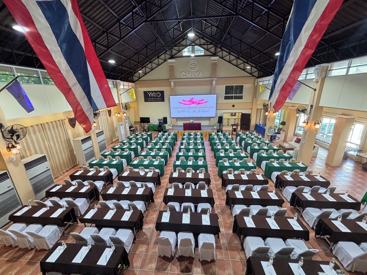 A conference room with rows of tables and chairs, a stage with a presentation screen, and flags hanging from the ceiling.