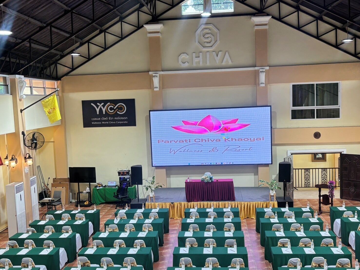Ein Konferenzraum mit Reihen von Tischen, einer Bühne mit einer großen Leinwand und einem Banner für Chiva Wellness & Resort.