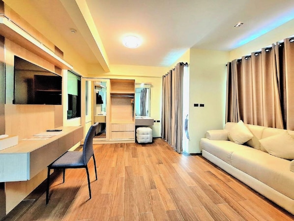 A modern hotel room with a wooden floor, a sofa, a desk with a chair, a TV, and a wardrobe. - Wellness World Khao Yai (Pak Chong)