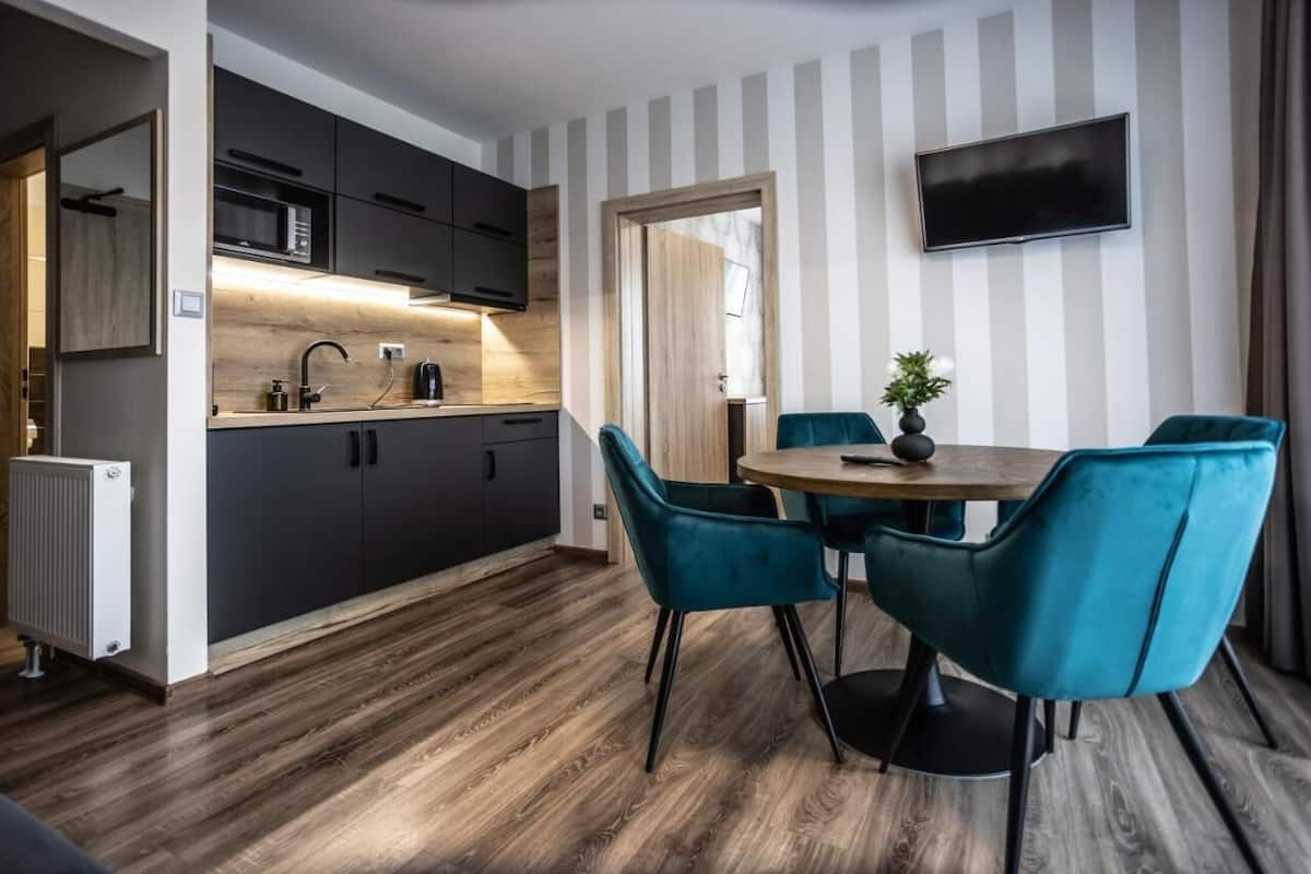 A modern kitchen with dark cabinets, a microwave, and a sink. There is a dining area with teal chairs and a small table. A TV is mounted on the wall, and a radiator is visible on the floor.