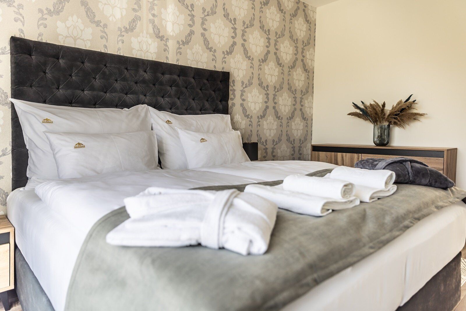 A neatly made bed with white linens and a grey throw blanket, a dark tufted headboard, and a nightstand with a vase of dried flowers.