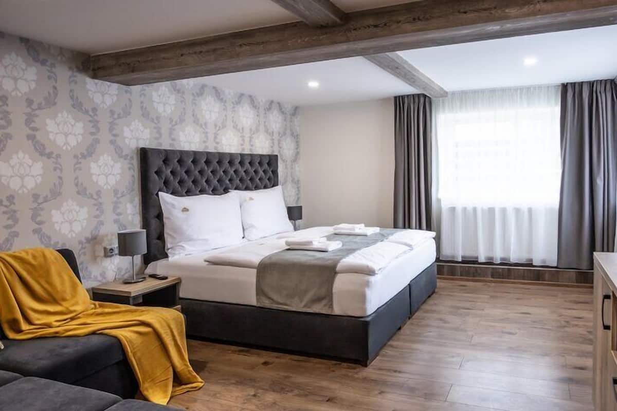 A modern bedroom with a large bed, wooden floor, and a nightstand with a lamp.