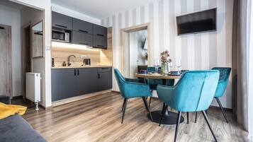 Una cocina moderna con armarios oscuros, encimera de madera y microondas empotrado. Hay una zona de comedor con una mesa, cuatro sillas azules y un jarrón con flores. Un televisor de pantalla plana está montado en la pared sobre la zona de comedor.