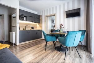 A modern kitchen with dark cabinets, a wooden countertop, and a built-in microwave. There is a dining area with a table, four blue chairs, and a vase with flowers. A flat-screen TV is mounted on the wall above the dining area.