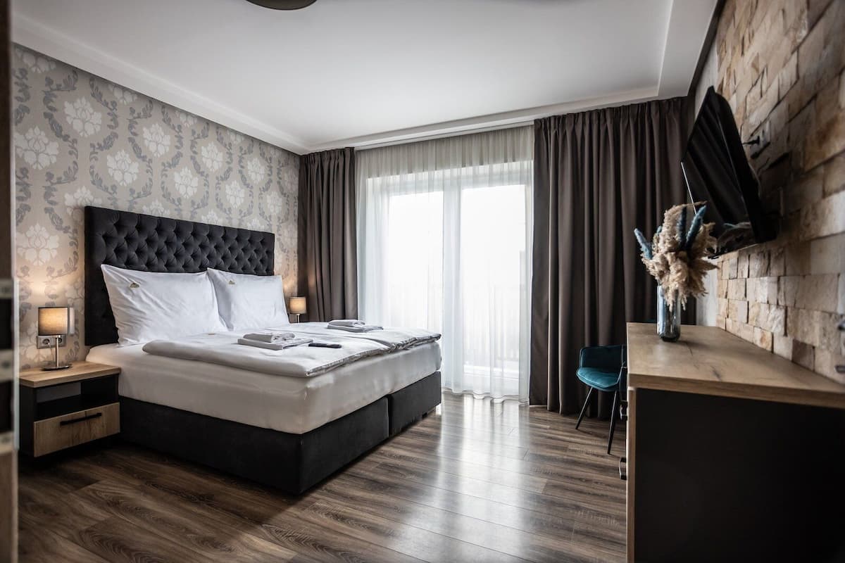 A modern hotel room with a tufted headboard bed, a wooden desk, a flat-screen TV, and a large window with curtains.