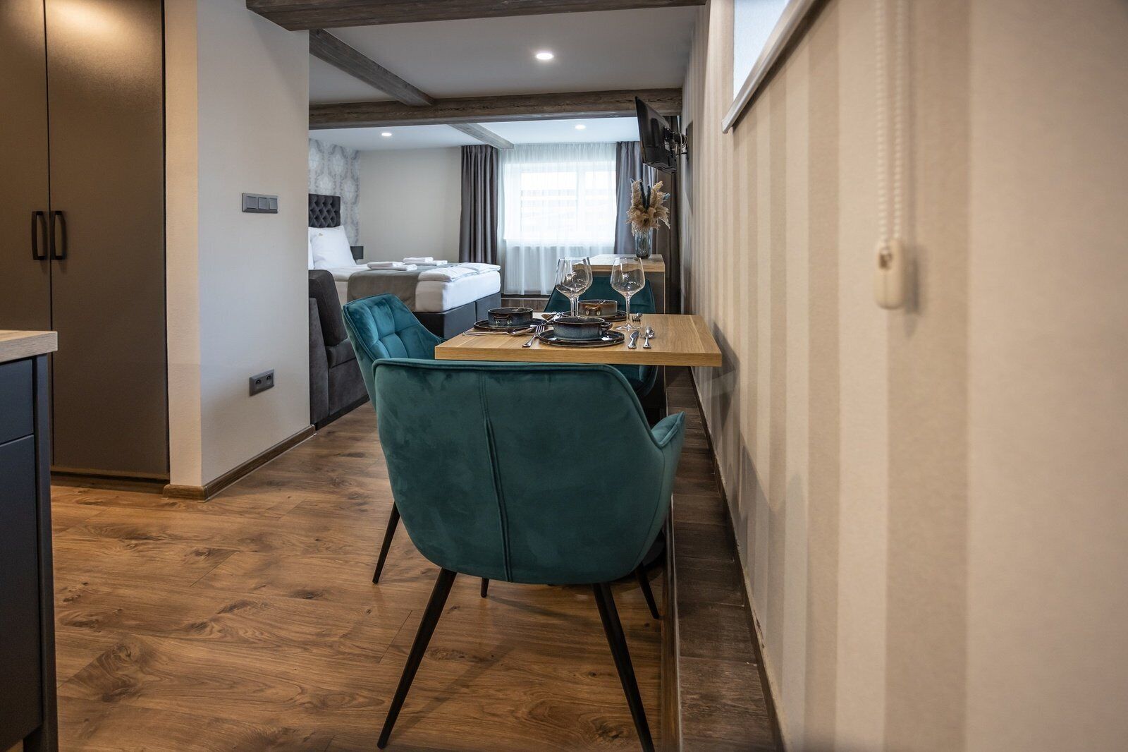 Ett modernt hotellrum med trägolv, turkosa stolar, ett trämatbord och en säng med en grå sänggavel.