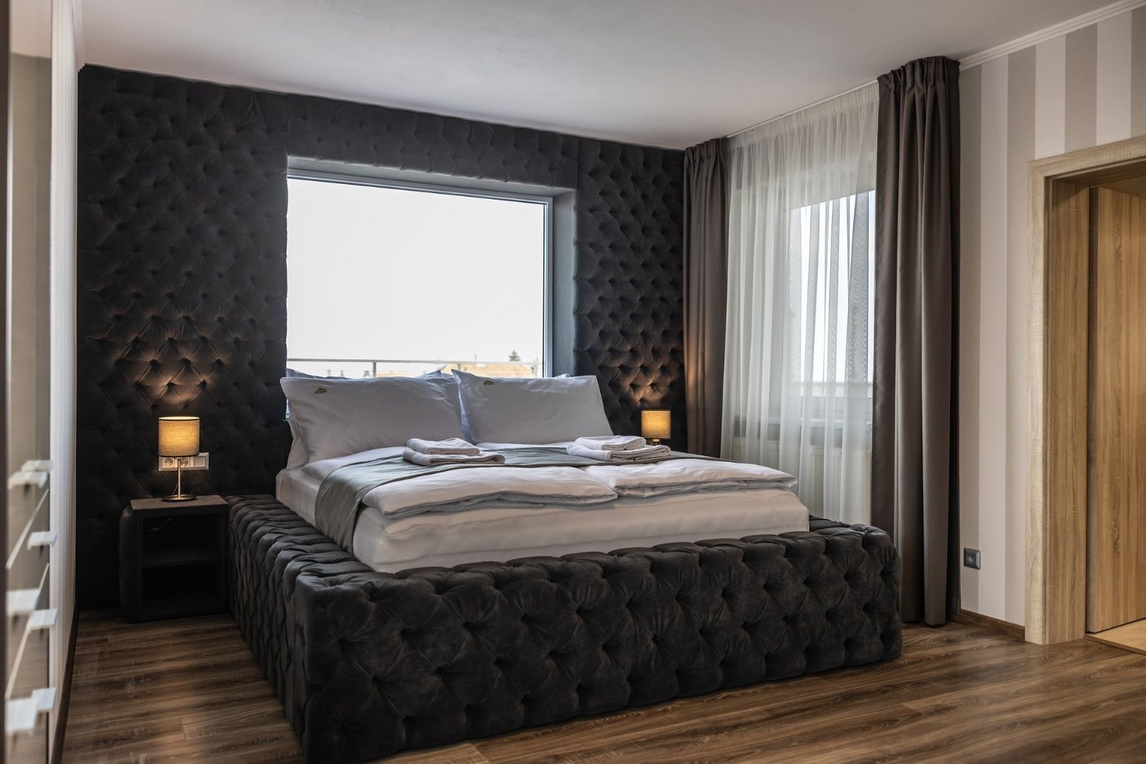 Un dormitorio moderno con una cama grande, paneles de pared oscuros, una ventana con cortinas y suelo de madera.