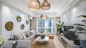 Una sala de estar moderna con un sofá, una mesa de centro y un televisor. Hay ventanas grandes con vistas a la ciudad, y la habitación tiene un diseño limpio y minimalista.