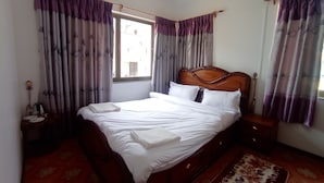 A neatly made bed with white linens, a wooden headboard, and a red patterned rug. There is a nightstand with a lamp and a small table with a kettle and cups. - Janaki House (Burhanilkantha)