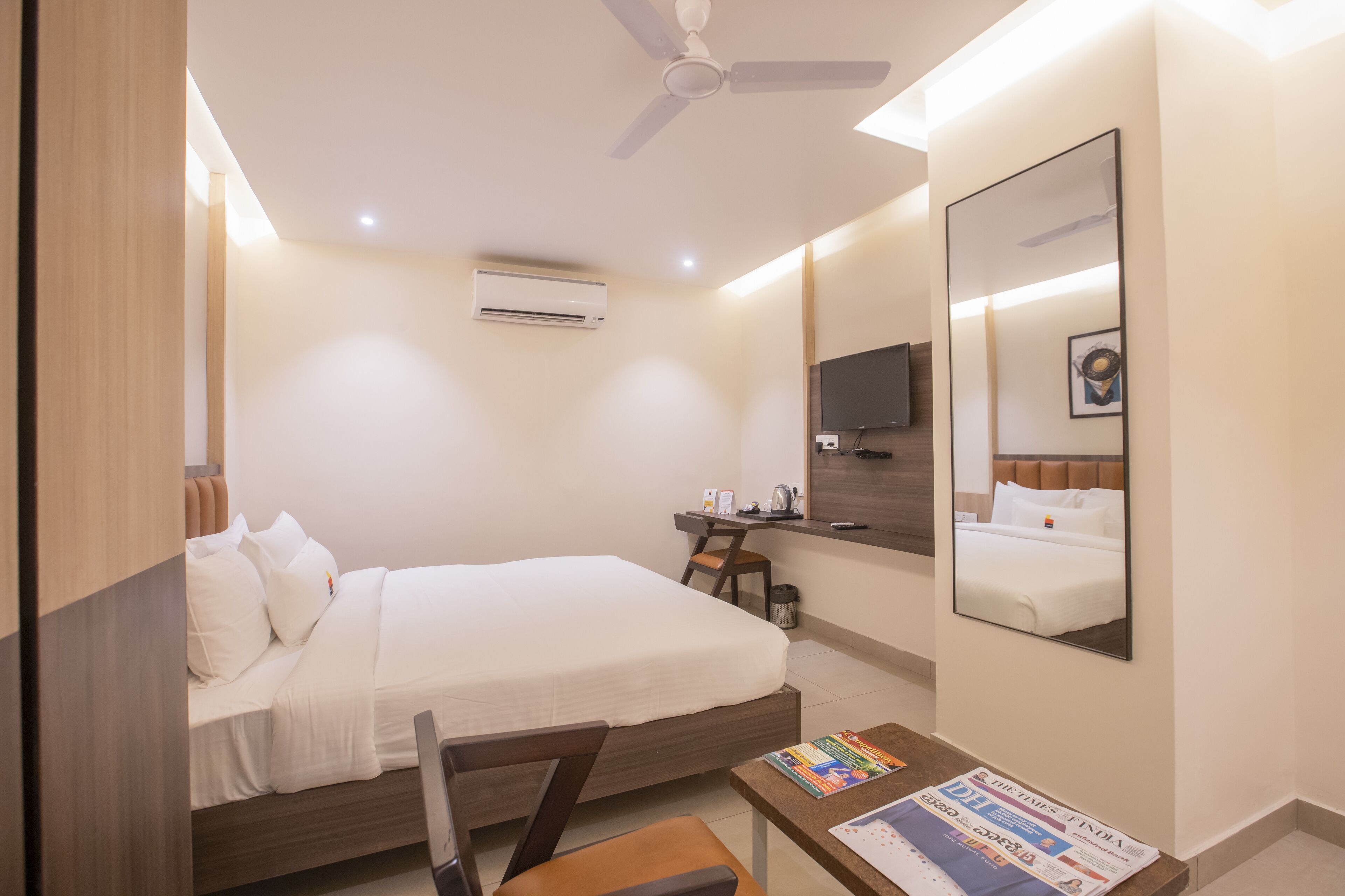 Habitación de hotel con cama, escritorio, silla, televisor, espejo y ventilador de techo.