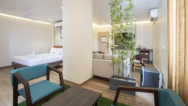 A modern hotel room with a bed, a sofa, a dining table, and a large potted plant.