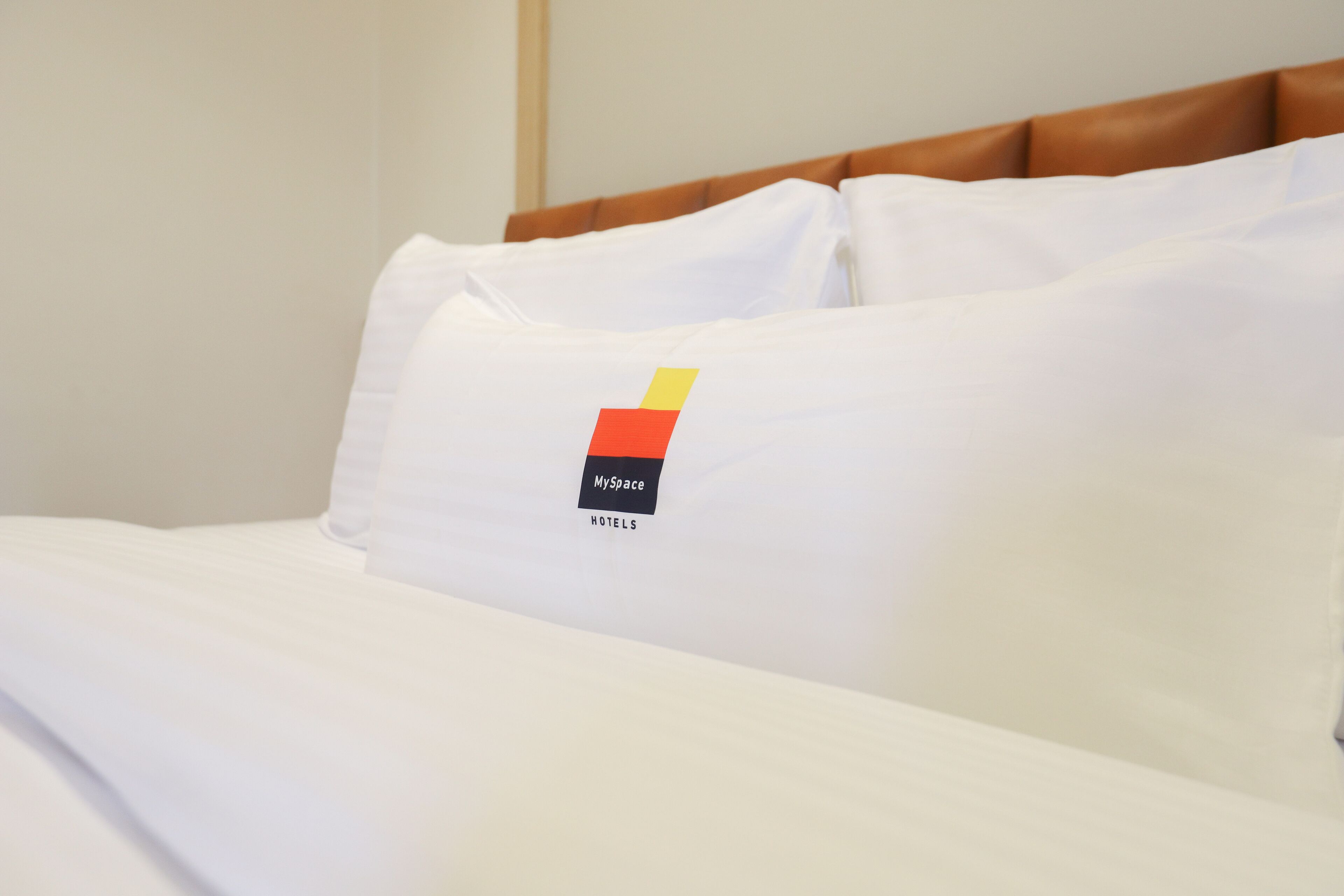 A hotel bed with white bedding and a pillow featuring a logo with the text "MySpace HOTELS".