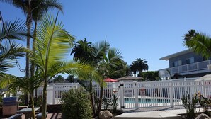 A pool area with a white fence, palm trees, and a building with a red roof in the background. - Ultimate Beach Vacation in the Heart of Carlsbad  (Carlsbad)