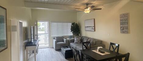 A spacious living area with a dining table, chairs, and a sofa. A ceiling fan is visible, and there is a window with blinds.