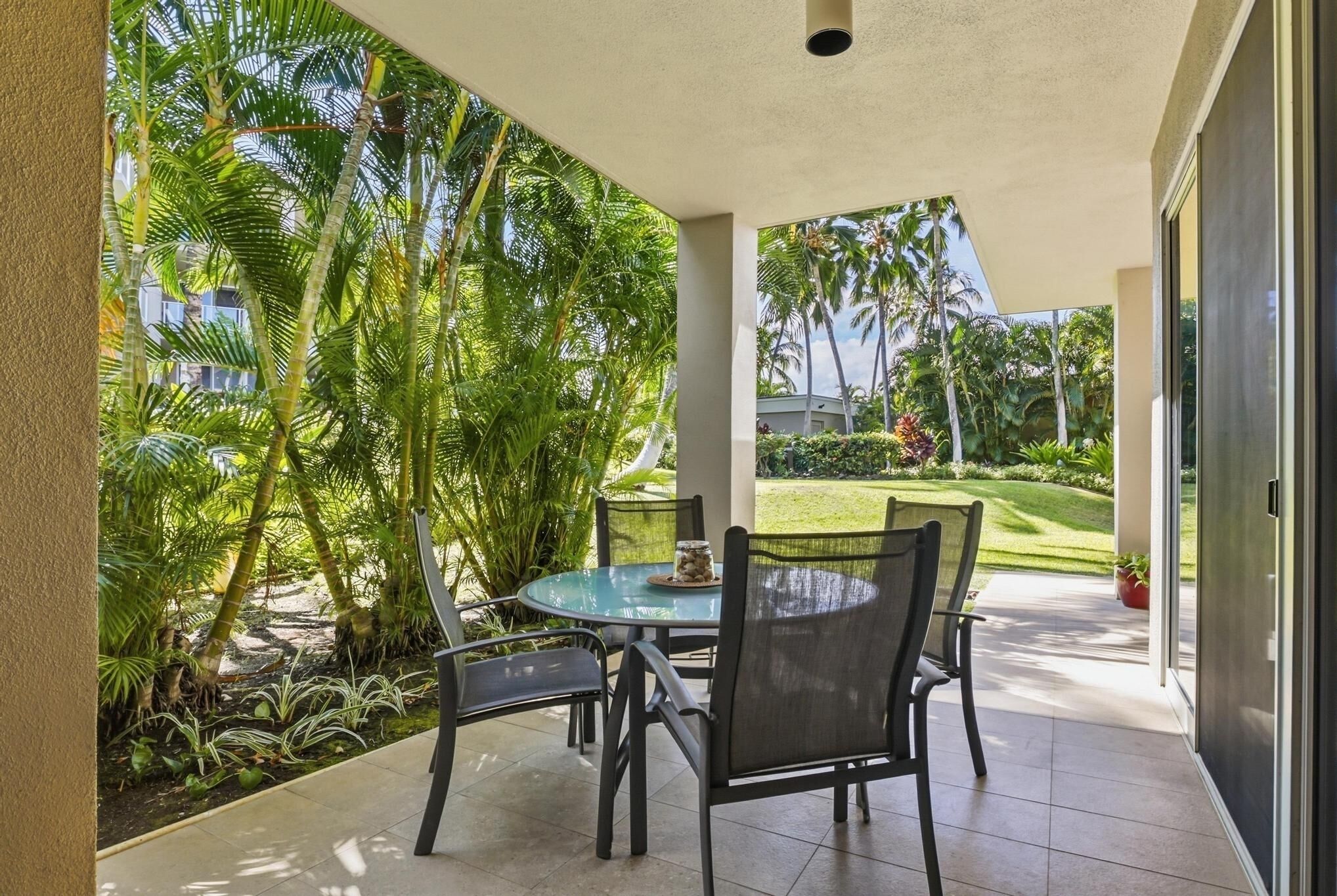A covered patio with a glass table and chairs, surrounded by lush tropical plants and a view of a well-maintained lawn and palm trees.