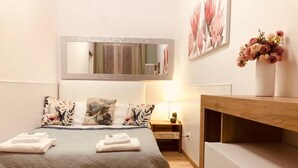 Et pænt opredt soveværelse med en seng, puder, et sengebord med en lampe, et spejl og en vase med blomster.
