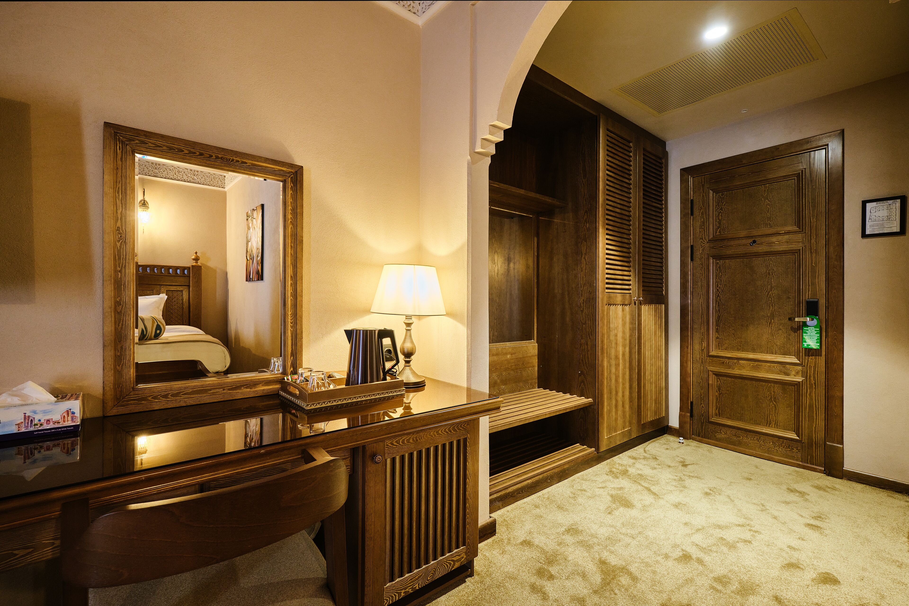 Hotelkamer met een houten bureau, een spiegel, een lamp, een bed en een deur met een groen bordje.