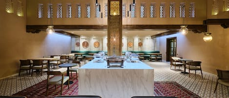 Un comedor amplio con una mesa buffet central, rodeado de sillas y mesas, bajo un techo alto con vigas de madera e iluminación decorativa.