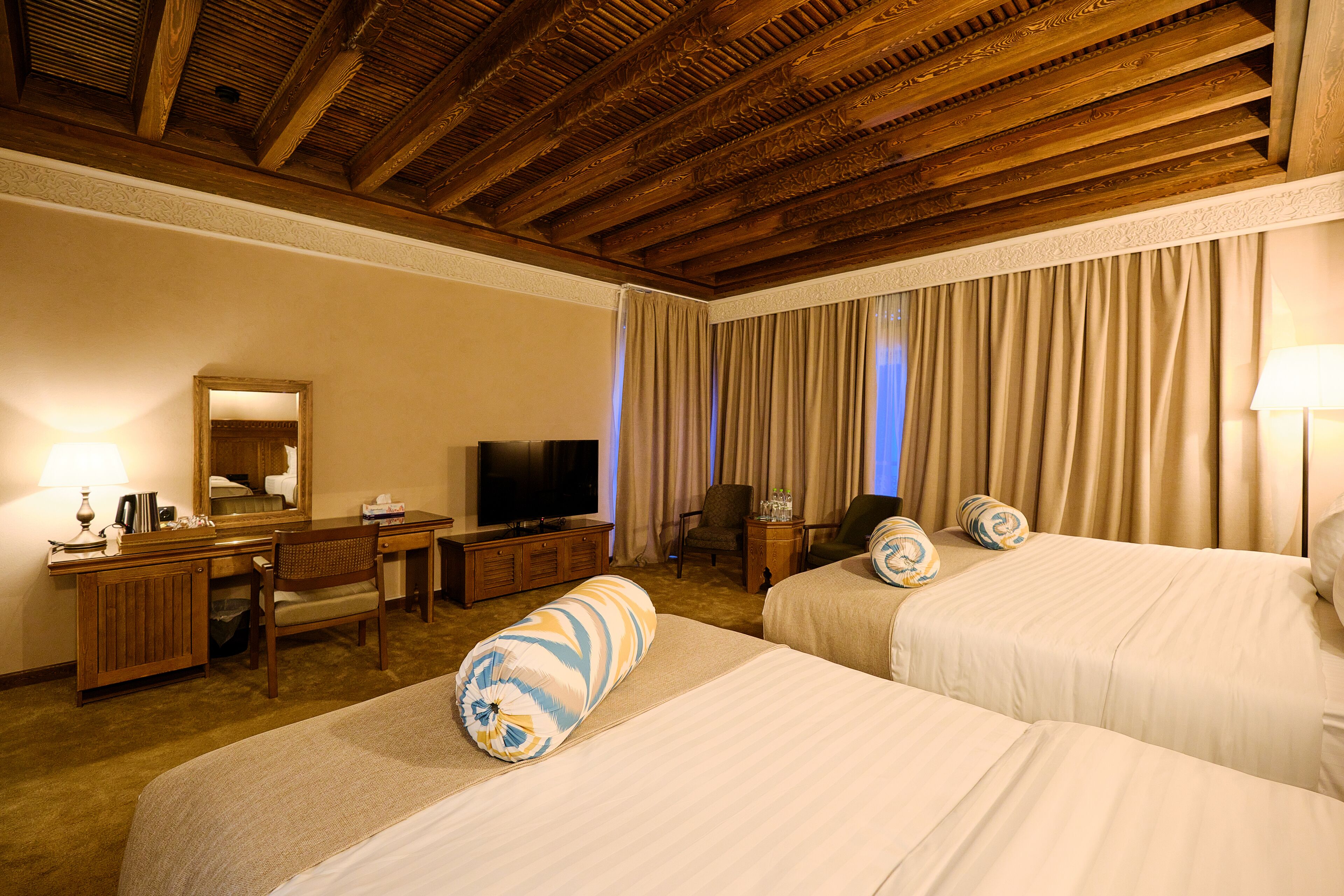 A hotel room with two beds, a desk, a television, and a large window with curtains.