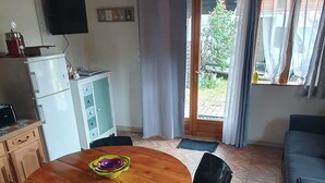 Een kamer met een houten plafond, een ronde houten tafel met stoelen, een televisie aan de muur, een kleine koelkast en een schuifdeur van glas die leidt naar een buitengebied.