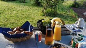 Een ontbijt buiten met een gedekte tafel voor twee personen, inclusief een broodmand, een schaal fruit, een juskan en een bord met eieren en toast.