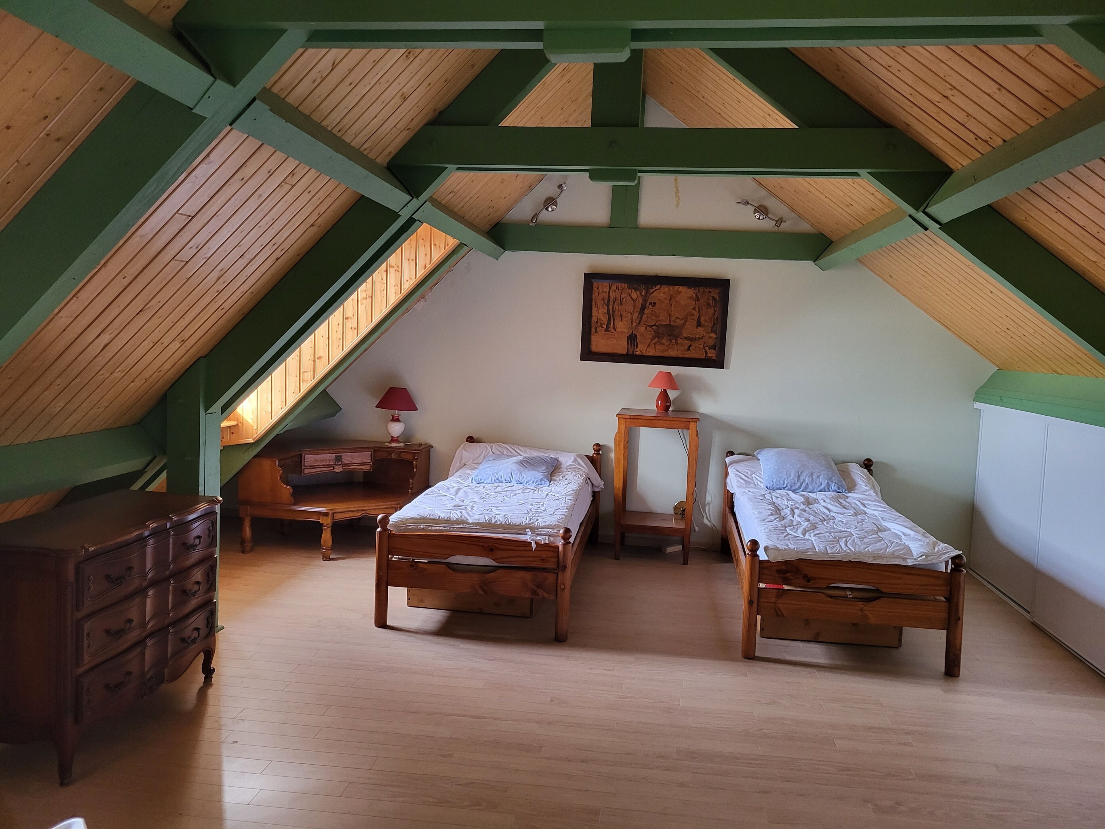 A room with a sloped ceiling, wooden beams, two wooden beds, a dresser, a nightstand, a lamp, and a painting on the wall.