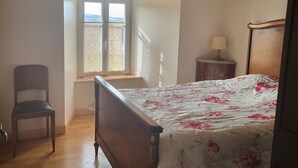 A bedroom with a wooden ceiling, a bed with a floral bedspread, a chair, a nightstand with a lamp, and a window with a view of the outdoors. - Proche Laguiole - Maison au Calme - vue Imprenable (Curières)