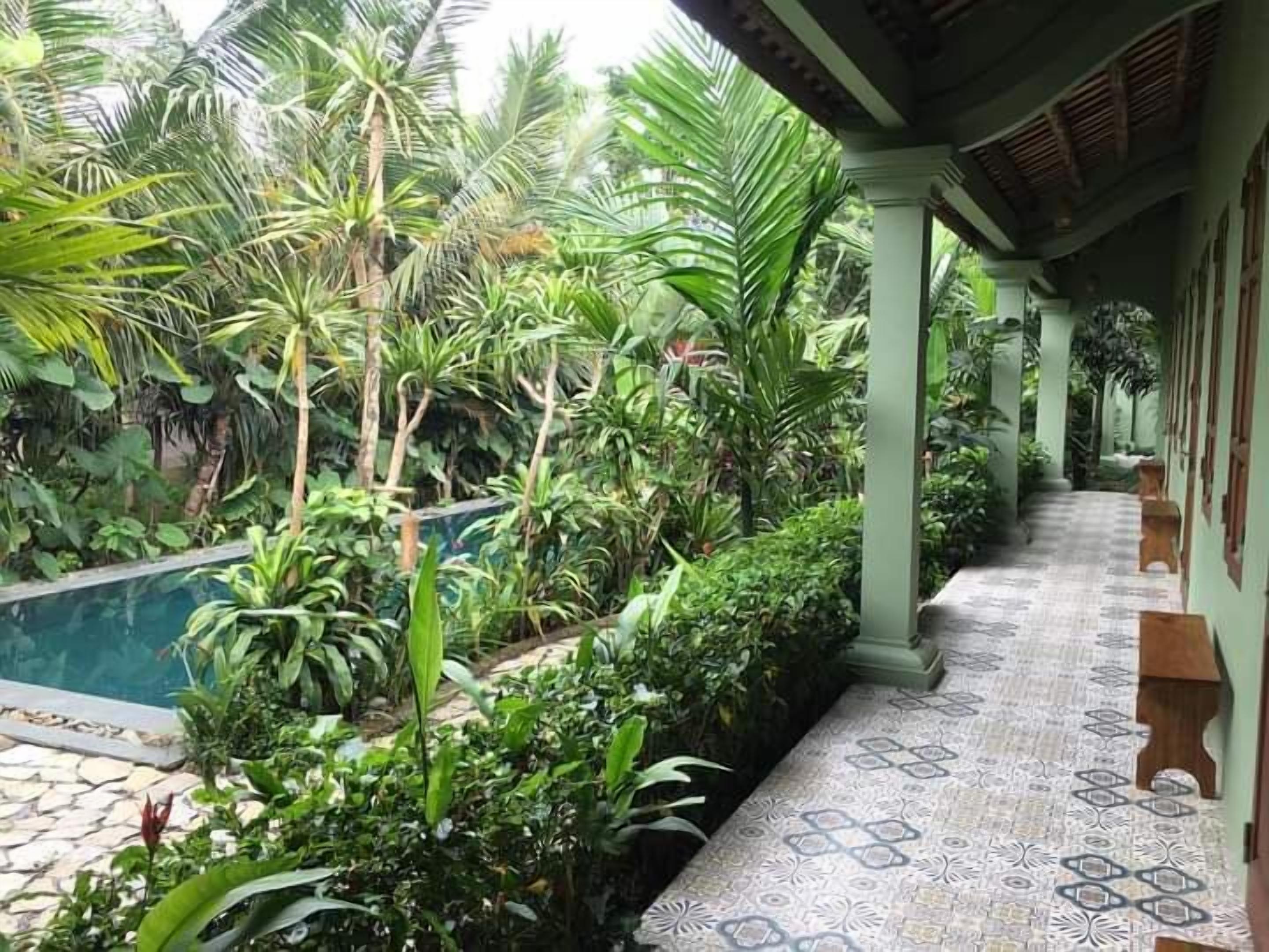 A poolside area with a tiled walkway, lush greenery, and a covered section with columns.