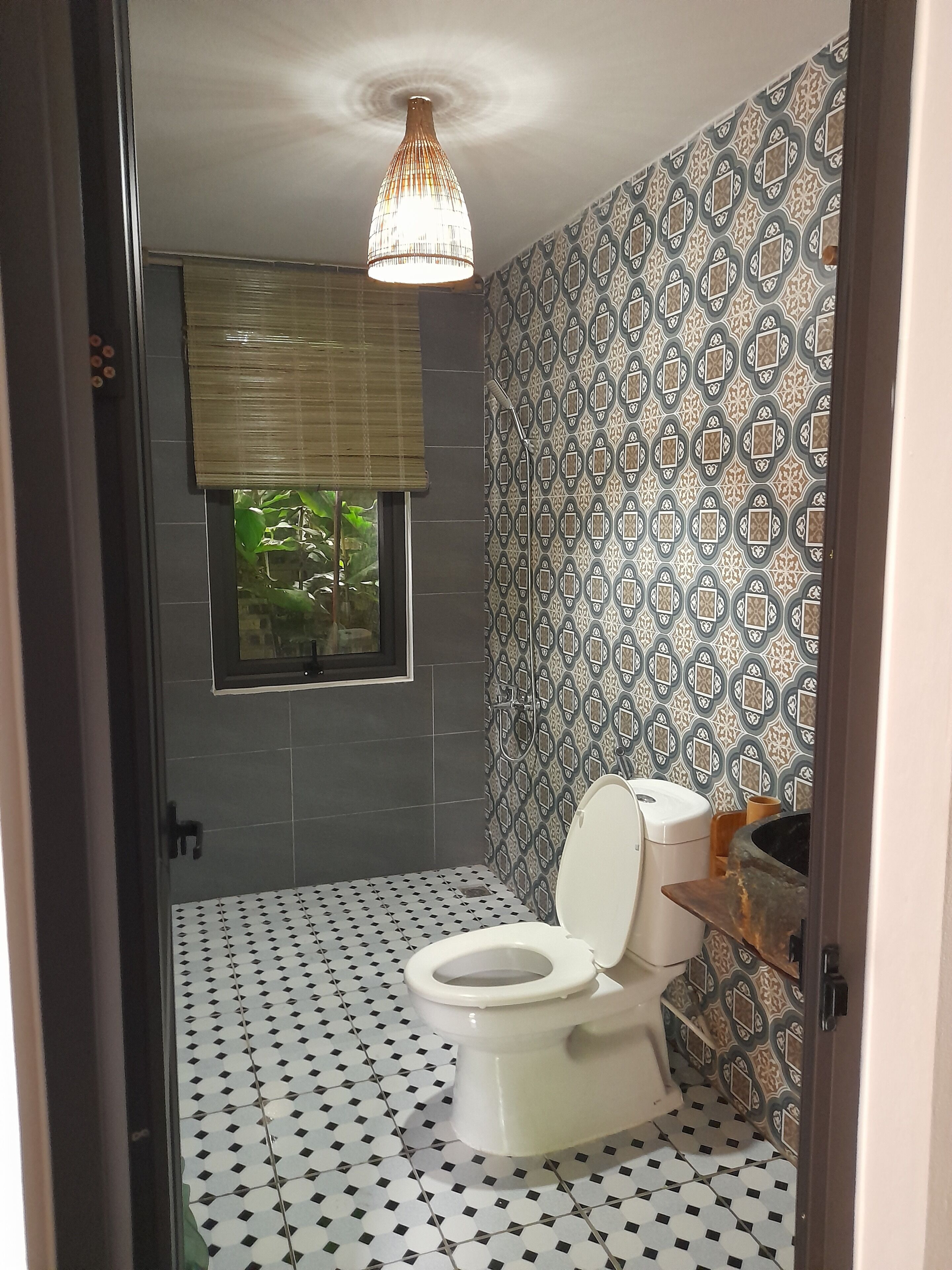 A bathroom with a patterned tile floor, a toilet, a window with a view of greenery, and a patterned wall.