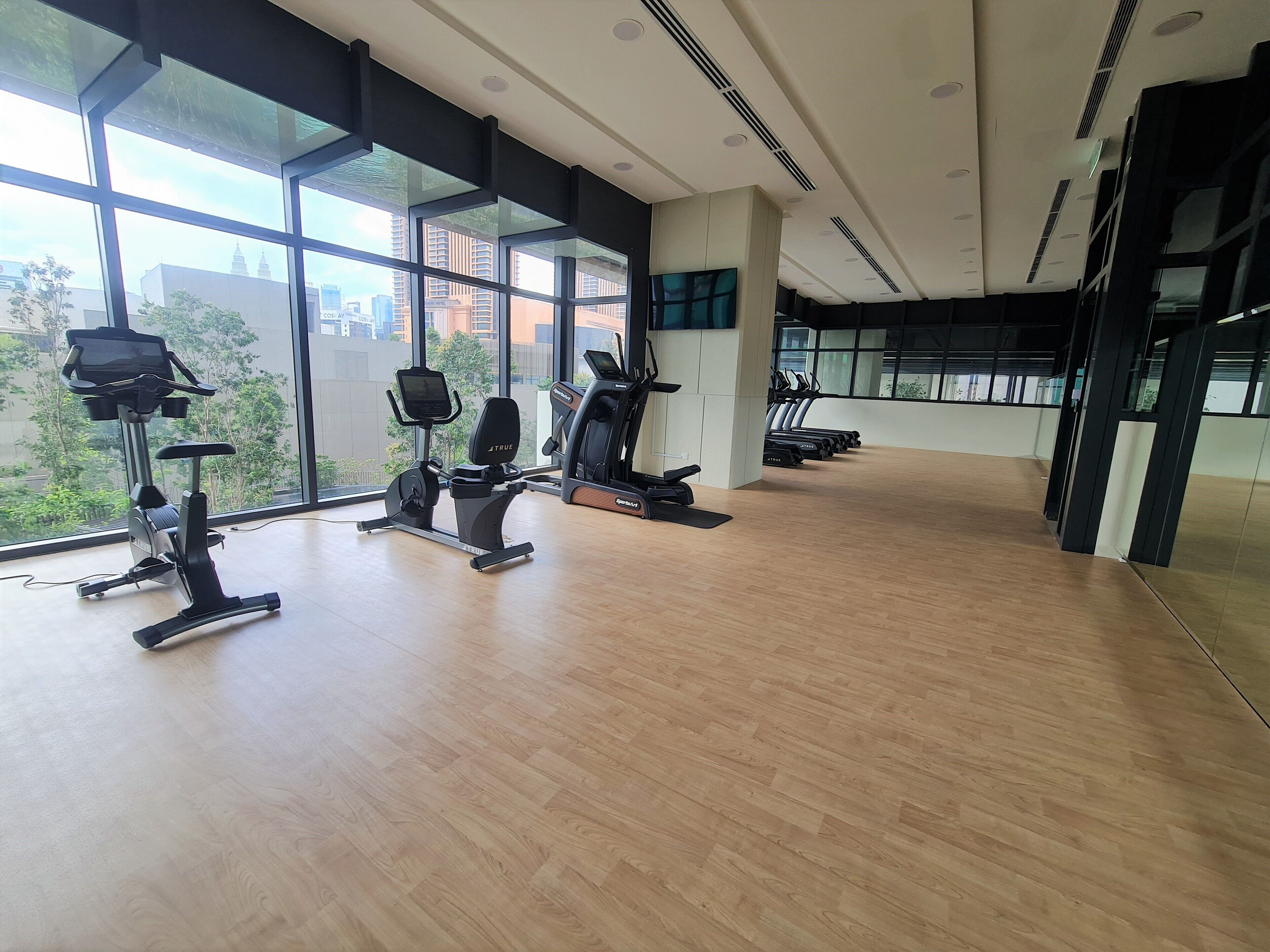A modern gym with cardio machines, large windows, and a wooden floor.
