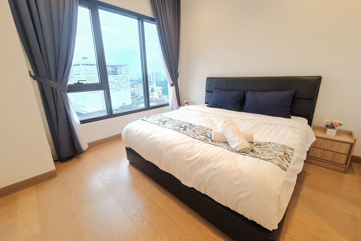 Een opgemaakt bed met een bedsprei en kussens met een patroon, een houten vloer, een nachtkastje met een bloemenvaas en een groot raam met gordijnen.
