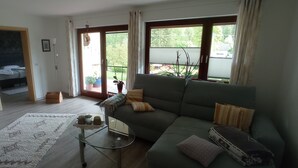 Uma sala de estar com um sofá verde, uma mesa de centro de vidro, um armário branco e vista para um jardim através de uma porta deslizante.