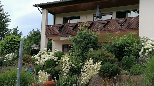 Um edifício de dois andares com telhado marrom, rodeado por vegetação exuberante e plantas floridas.