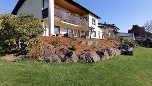 Uma casa moderna de dois andares com varanda, cercada por um jardim bem cuidado e pedras grandes.