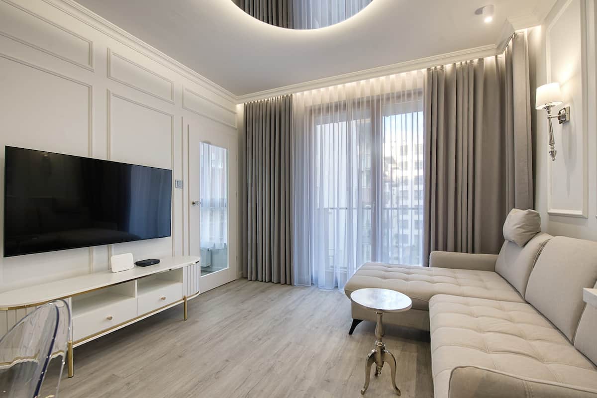 A modern living room with a flat-screen TV, a beige sofa, a small round table, and a large window with curtains.