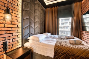 Une chambre d’hôtel moderne avec un mur en briques, un grand lit avec une literie blanche, une couverture matelassée marron et une vue sur la ville par la fenêtre.