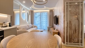 A modern hotel room with a round wooden dining table, a sofa, a TV mounted on the wall, and a large window with curtains.