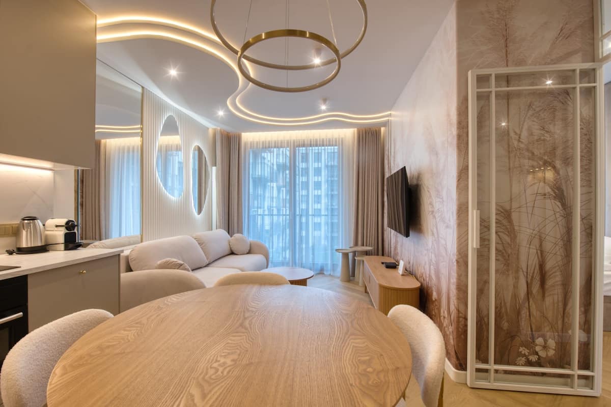 A modern hotel room with a round wooden dining table, a sofa, a TV mounted on the wall, and a large window with curtains.