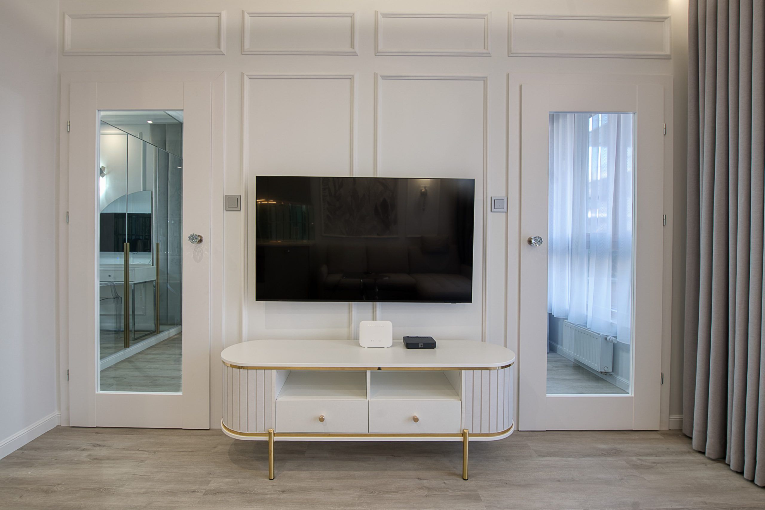 Una sala de estar moderna con un televisor de pantalla plana montado sobre un mueble blanco con patas doradas, una pequeña mesa blanca y un puerta de cristal que da a un balcón.